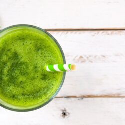 Tropical Crush Green Smoothie with paper white and green stripped straw mint leave on white wood table two more green and white stripped straws on table.