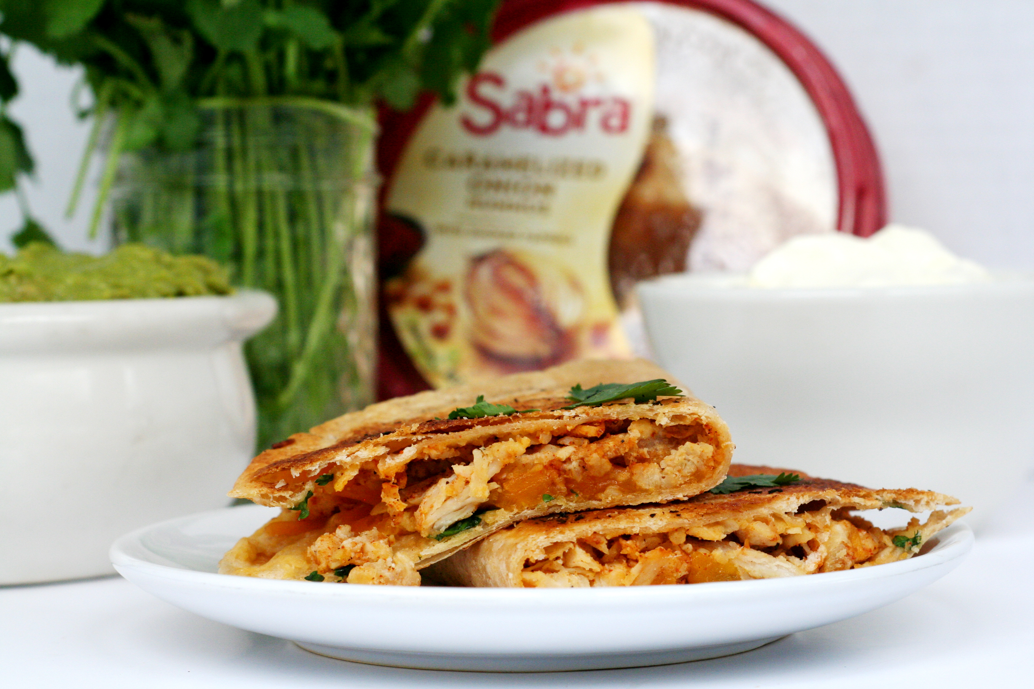 Sabra Hummus Chicken filled Quesadilla on white plate, cilantro, guacamole, sabra hummus and sour cream in the background.